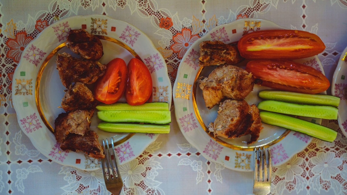 Two plates of grilled meat with sliced tomatoes and cucumbers on a patterned tablecloth